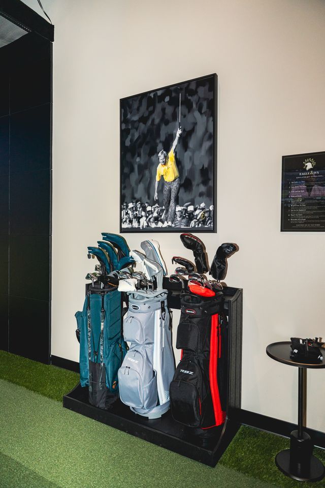 Three golf bags on a stand in a bay, under a framed print of a golfer celebrating a putt